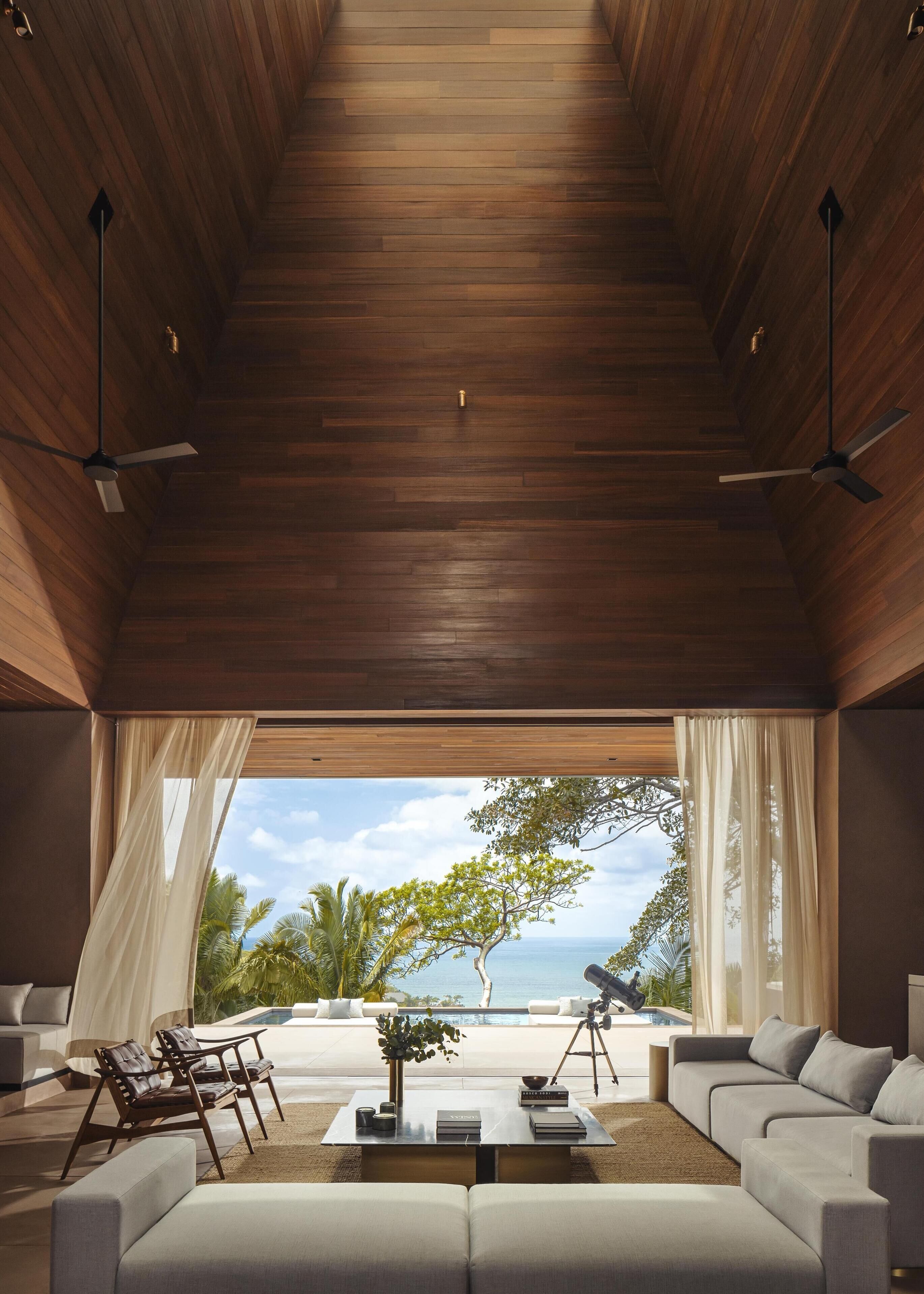 A modern living room with a high ceiling, wooden flooring, and large windows offering a view of the ocean. There are two sofas, a coffee table, and a telescope on a tripod.
