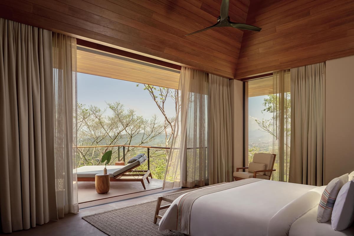 Un dormitorio con una cama grande, una silla, una mesita y vistas a árboles a través de una ventana.