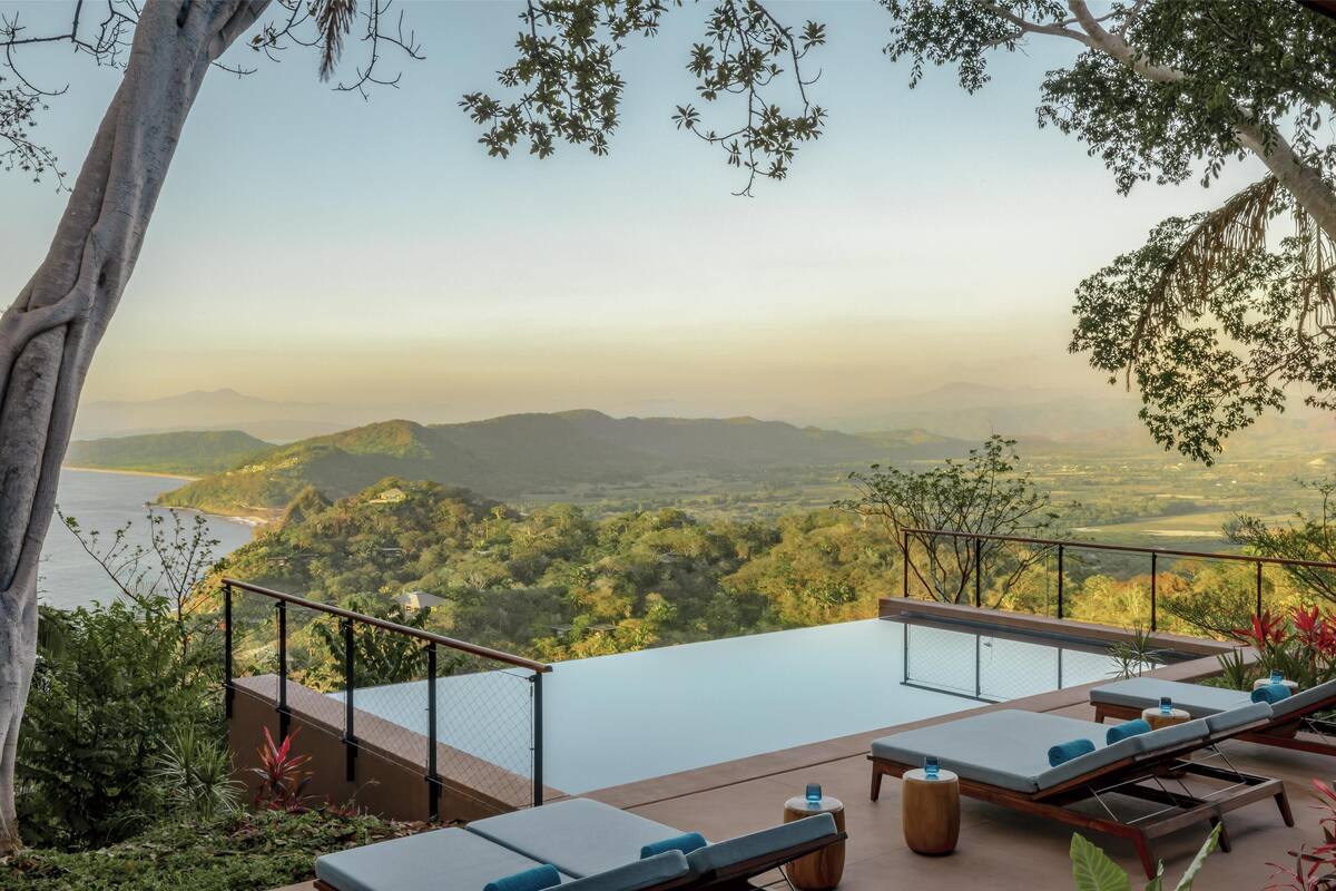A rooftop infinity pool with lounge chairs, surrounded by lush greenery and a scenic view of the ocean and mountains.