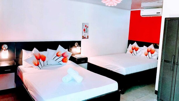 Ein Hotelzimmer mit zwei Betten, jeweils mit weißer Bettwäsche und roten und weißen Kissen. Es gibt einen Deckenventilator, eine Wandklimaanlage und einen kleinen Tisch mit einer Lampe.
