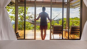 A person standing in a room with large glass doors, looking out at a wooden deck and outdoor furniture. - Cielo Lodge (Golfito)