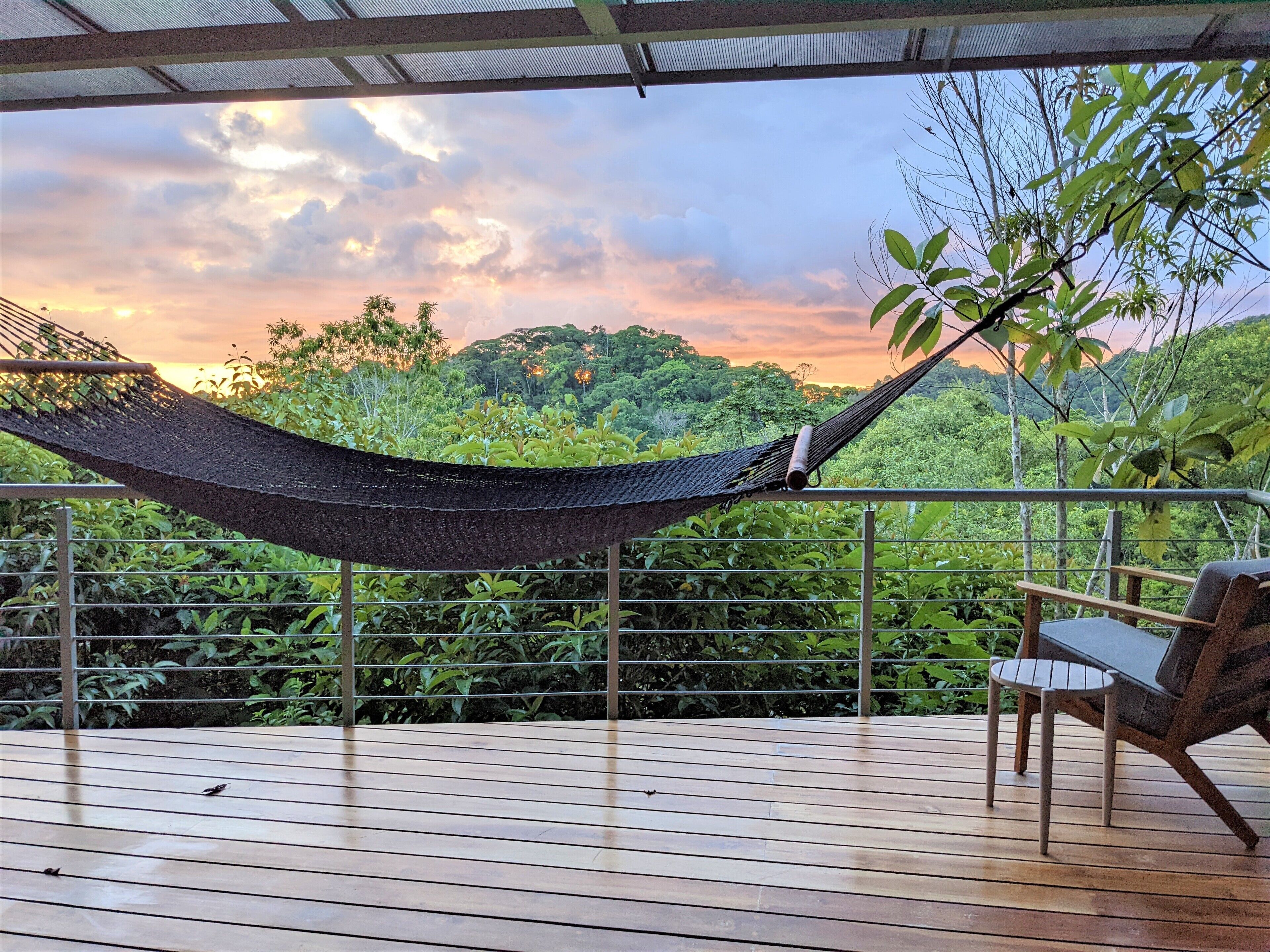 A hammock on a wooden deck with a view of lush greenery and a sunset.
