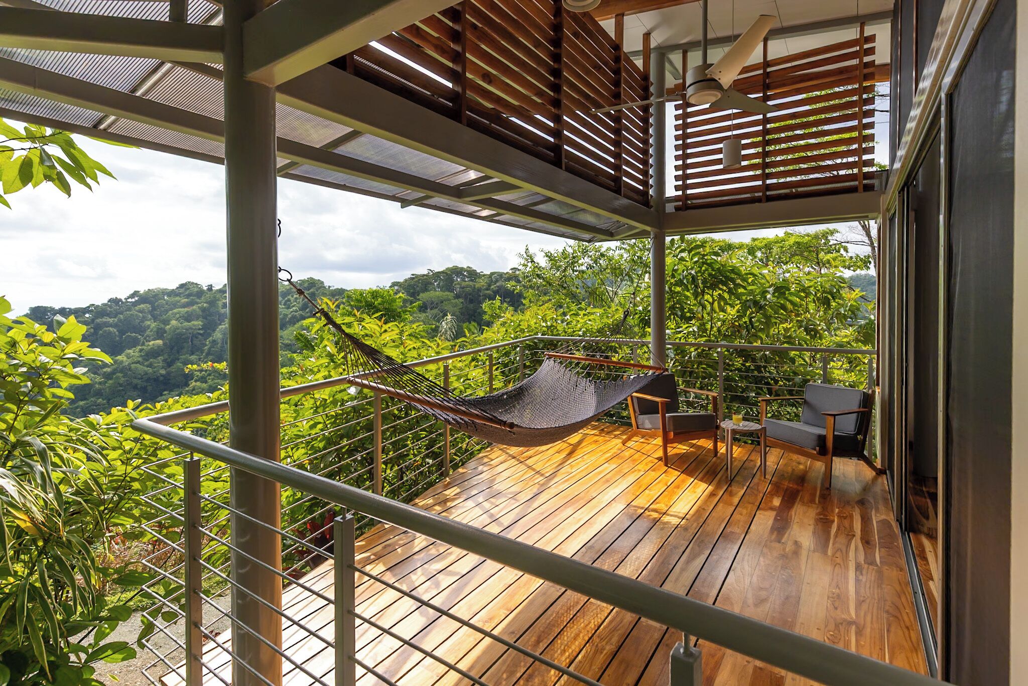 A wooden deck with a hammock, a table, and a chair, overlooking a lush green forest.