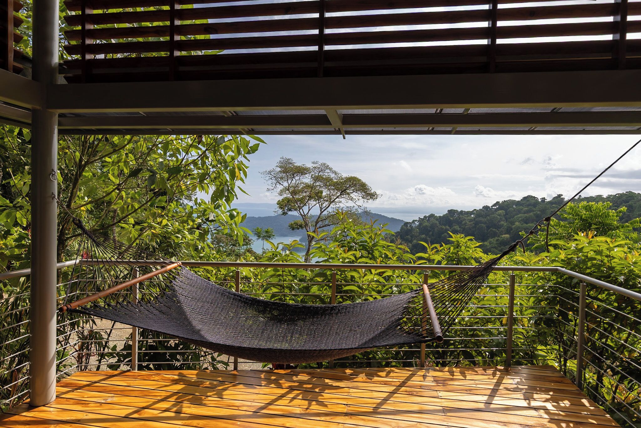 Un hamac sur une terrasse en bois, offrant une vue sur un paysage luxuriant et une étendue d’eau au loin.