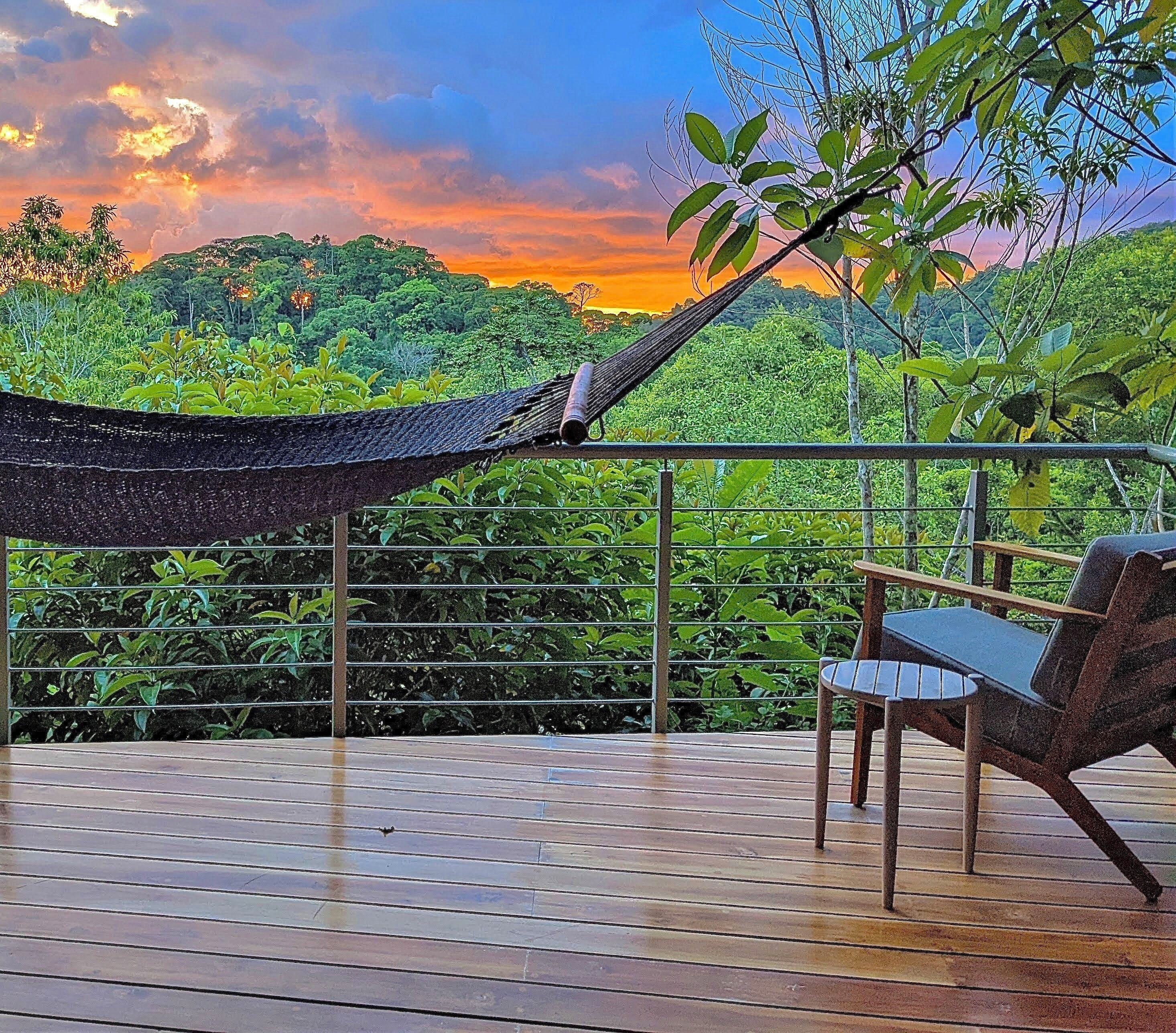 A hammock on a wooden deck with a chair, overlooking a lush green landscape and a sunset.