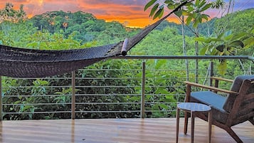 A hammock on a wooden deck with a chair, overlooking a lush green landscape and a sunset.