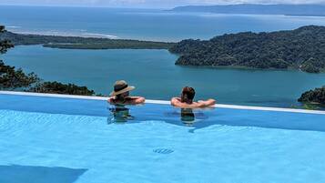 Relax by the infinity pool overlooking the ocean and distant islands, surrounded by lush greenery.