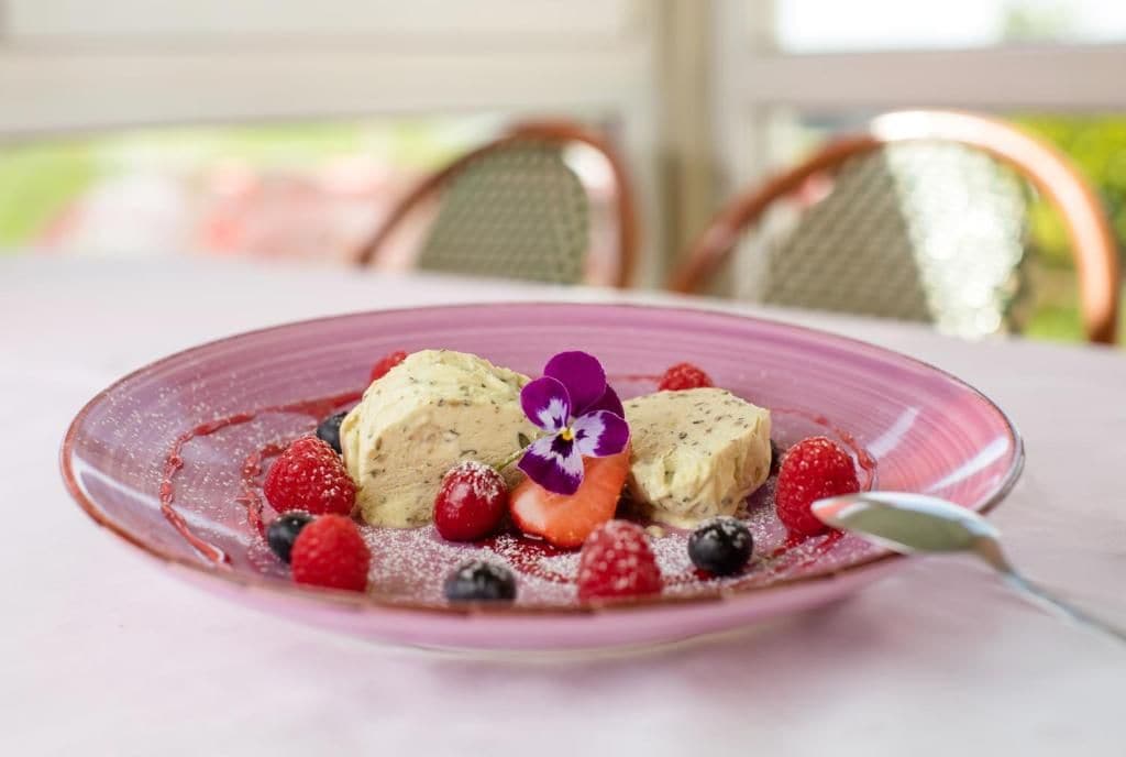 A dessert on a pink plate with berries and a flower, set on a table with chairs in the background.