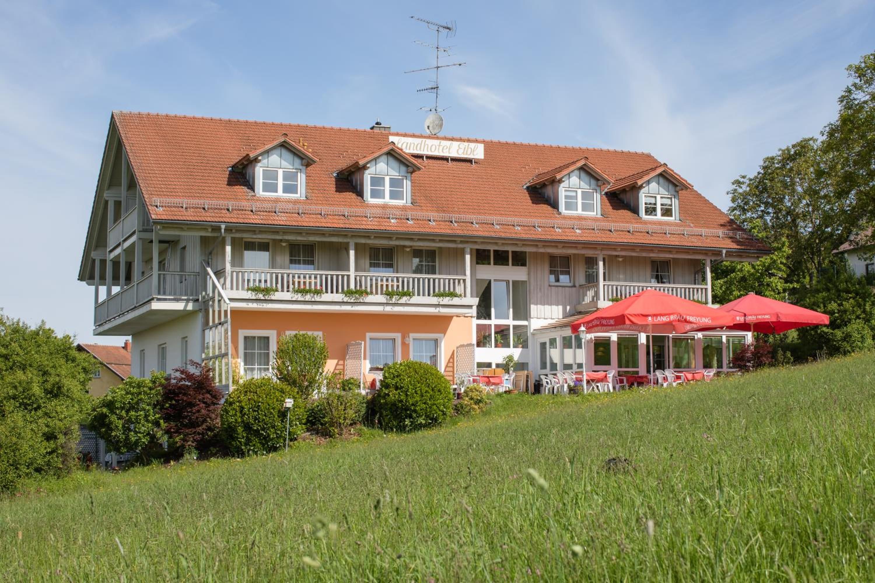 Un bâtiment à deux étages, doté d’un toit recouvert de tuiles rouges, d’un balcon et d’un espace extérieur aménagé avec des parasols rouges.