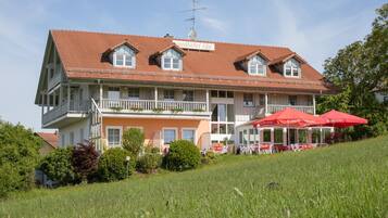 Un bâtiment à deux étages, doté d’un toit recouvert de tuiles rouges, d’un balcon et d’un espace extérieur aménagé avec des parasols rouges.