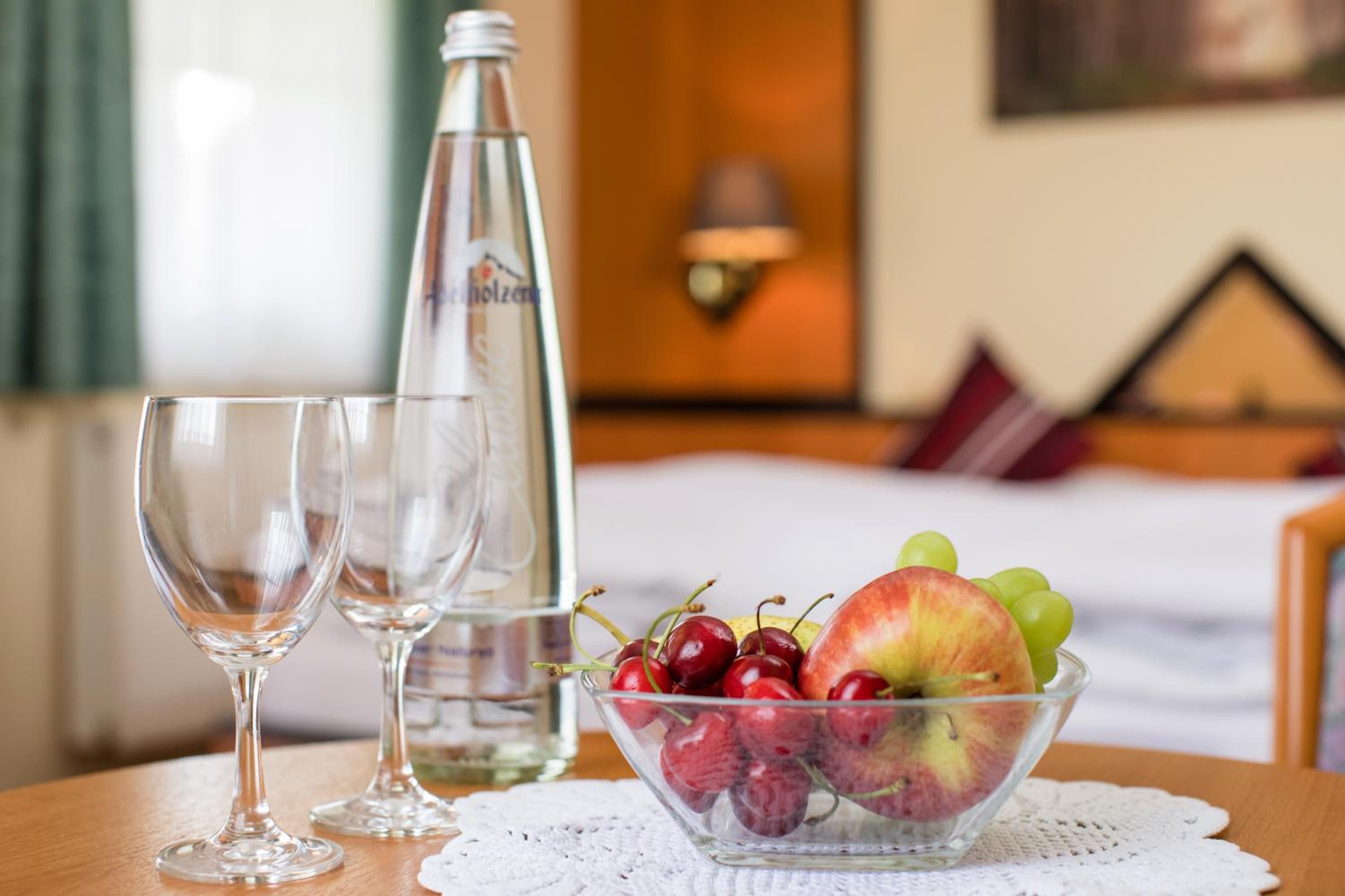 A hotel room with a bed, a bedside table with a bottle and glasses, and a bowl of fruit.