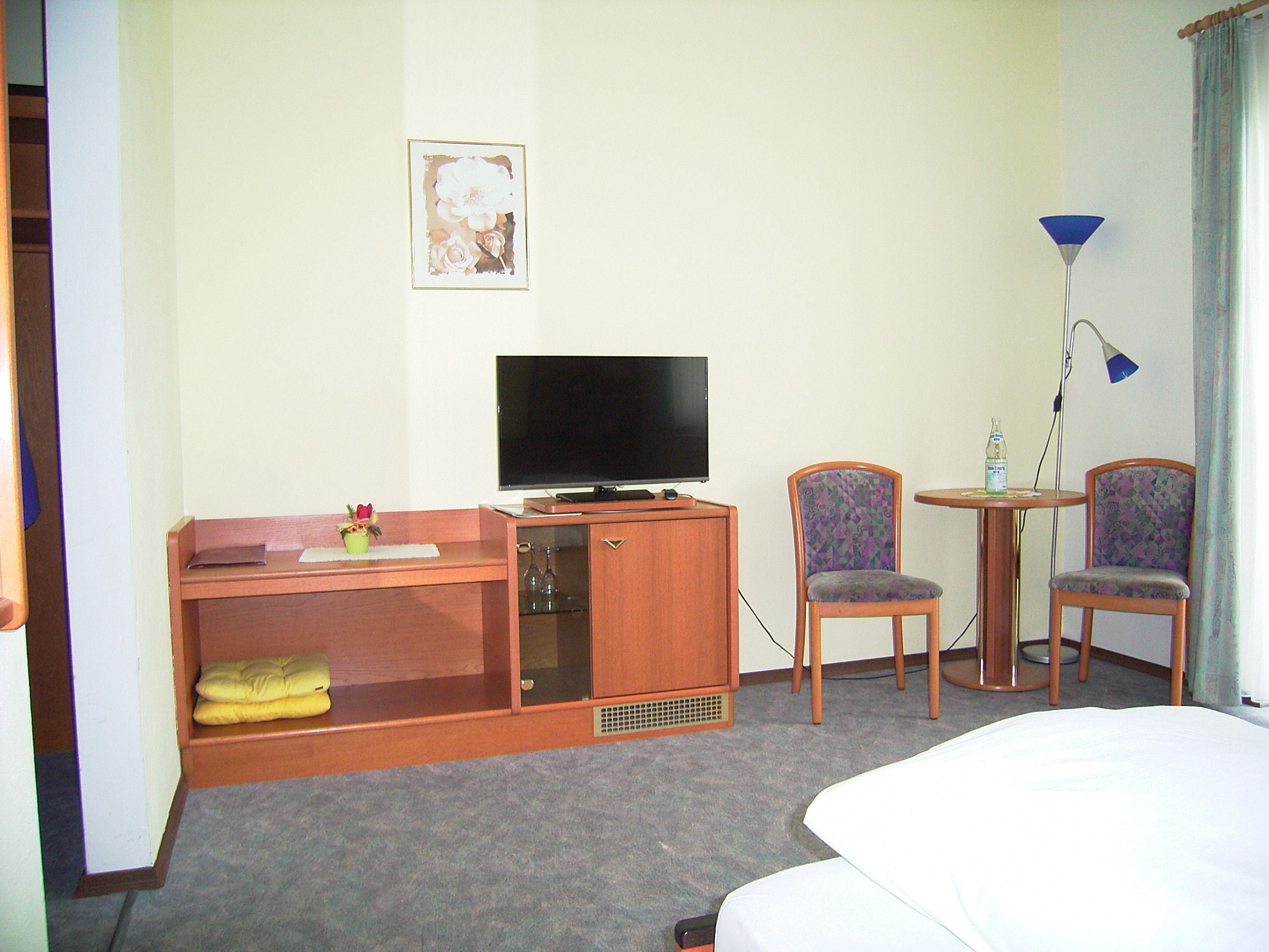 A hotel room with a bed, a desk with a television, two chairs, a small table with a bottle, and a lamp.