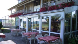 Un patio exterior con mesas y sillas, ventanas amplias y un balcón lleno de flores.