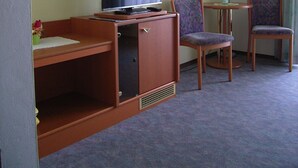 Habitación de hotel con cama, escritorio con televisor, dos sillas, una mesita, lámpara y un cuadro en la pared.