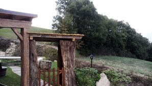 Un arc en bois portant l’inscription « PISCINA-POOL » mène à un espace piscine, entouré d’un chemin de gravier et de verdure.