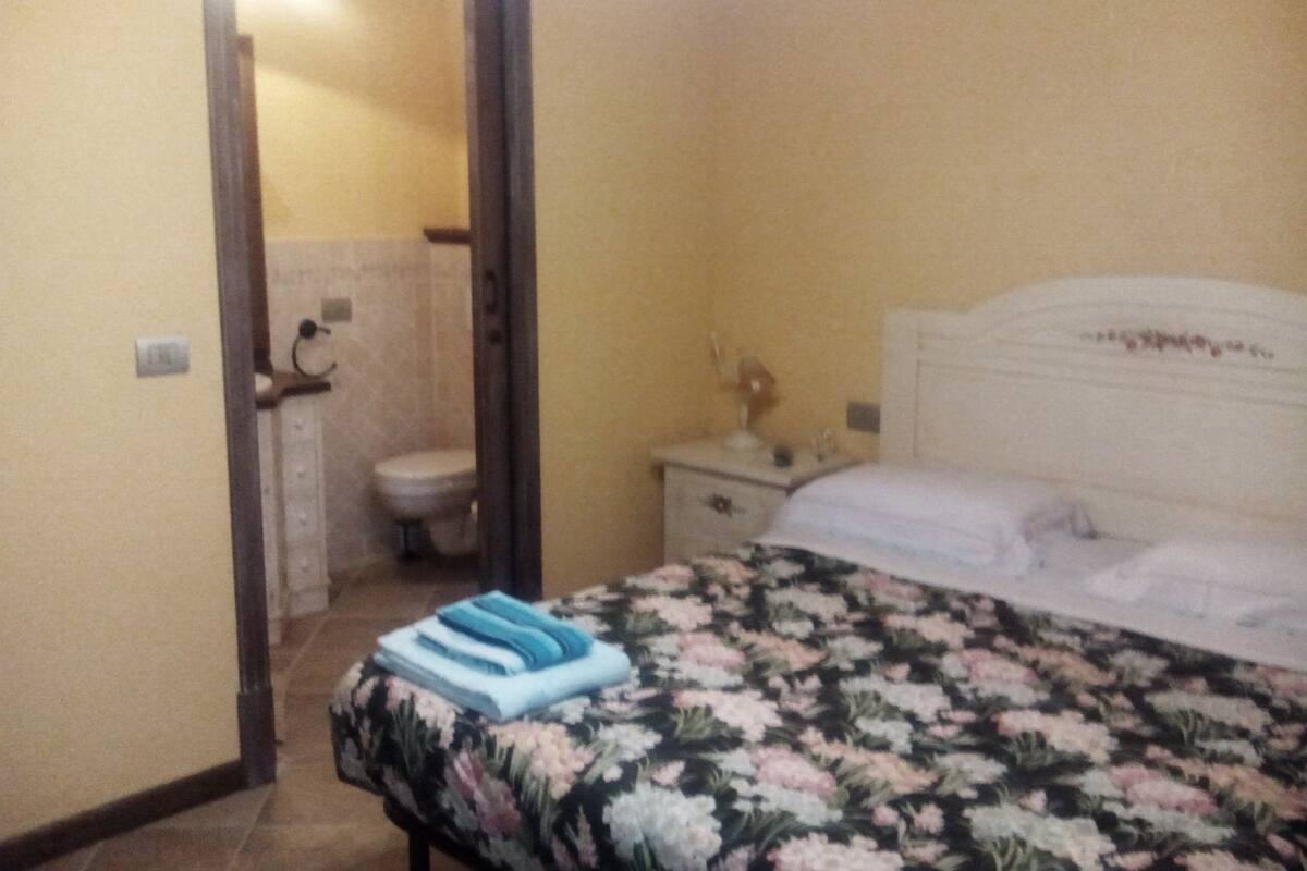 Ein Schlafzimmer mit Bett, Nachttisch und einem durchgehenden Türspalte sichtbarem Badezimmer.