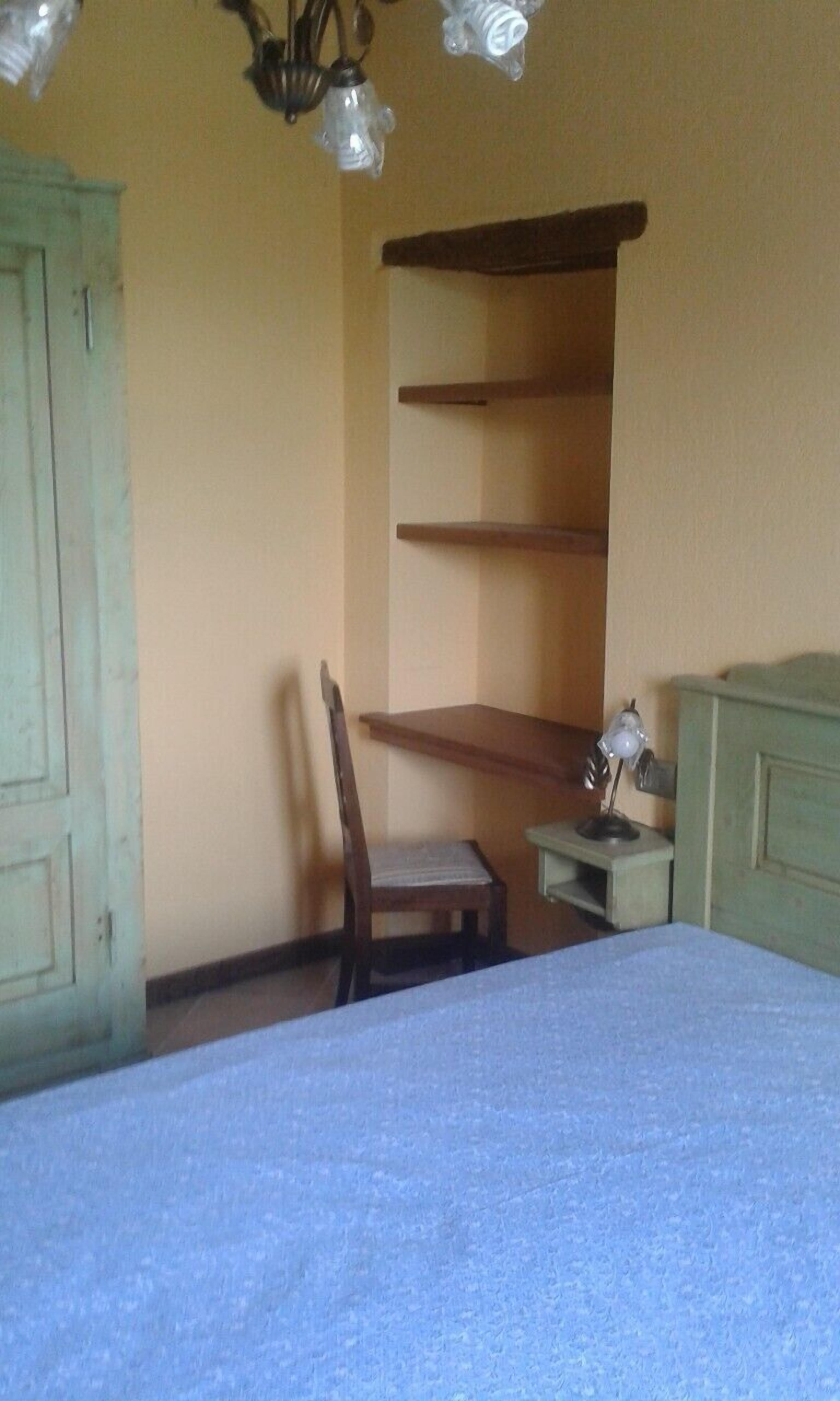 A room with a bed, a chair, a small table, and a shelf unit against the wall.
