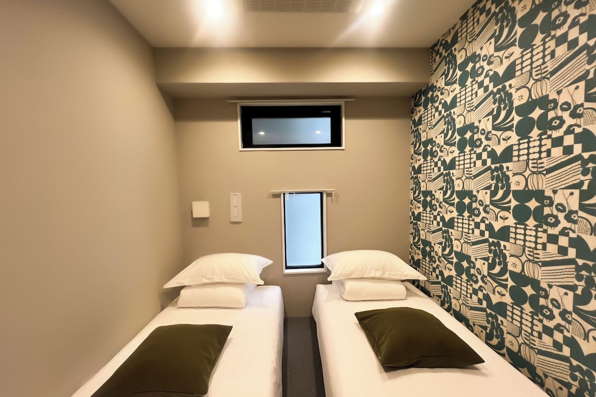 Una habitación pequeña y luminosa con dos camas individuales, una ventana y un papel pintado decorativo.