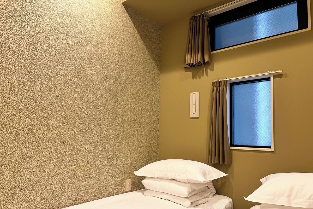 Una cama individual con ropa de cama blanca, un enchufe en la pared y una ventana con cortinas.