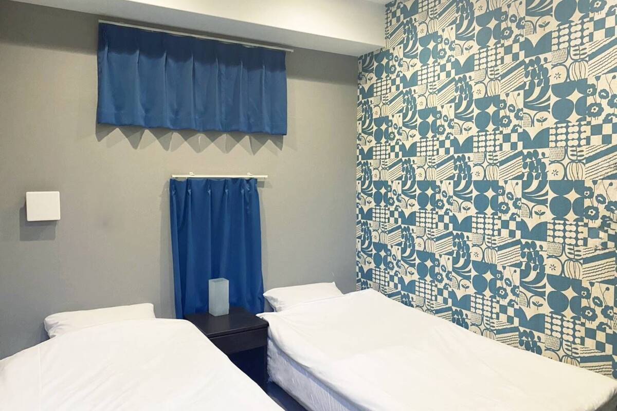 Habitación de hotel con dos camas, una cortina azul, un papel tapiz estampado y una mesita con lámpara.