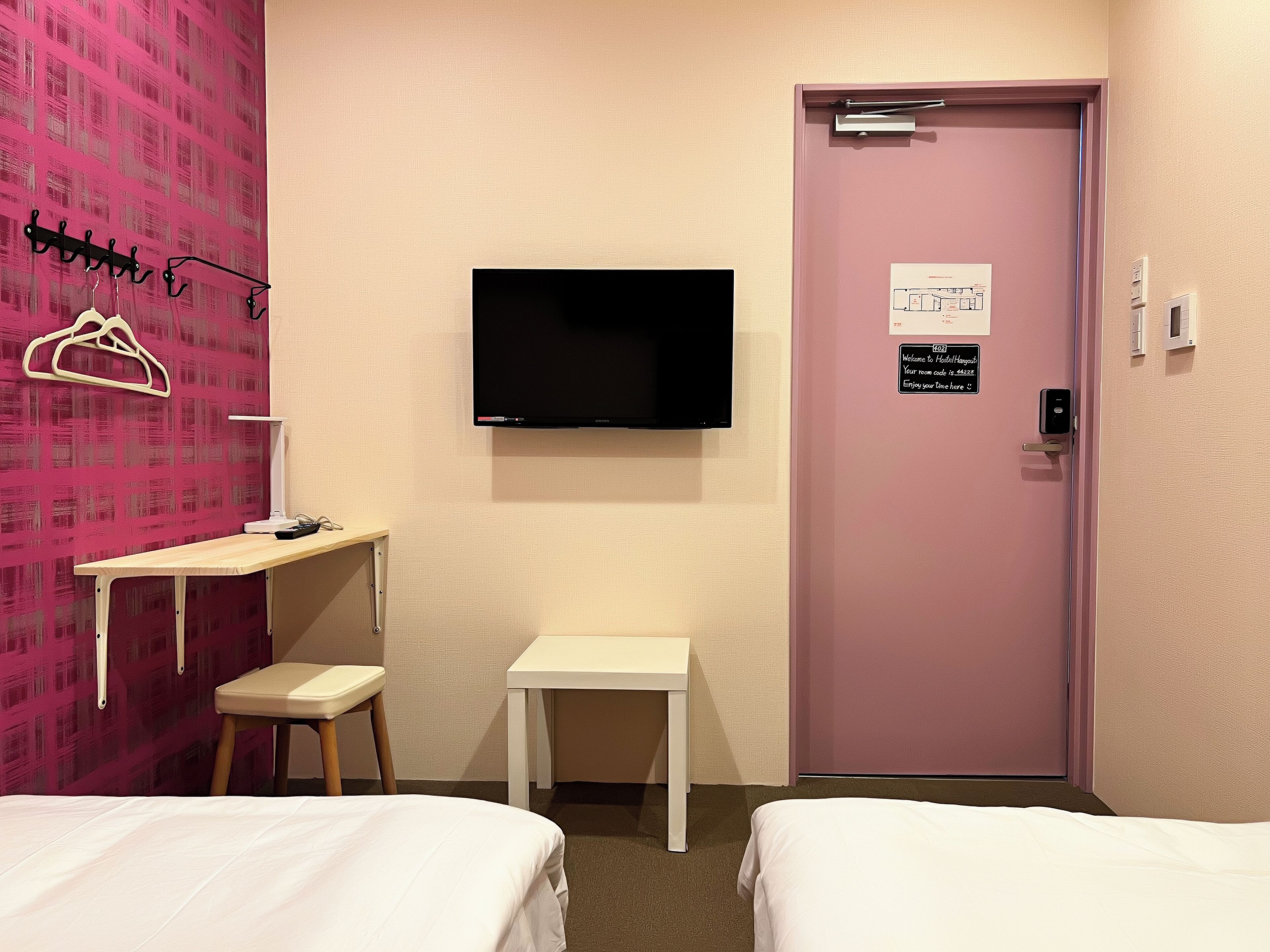 A hotel room with two beds, a desk, a chair, a TV, and a door with a sign.