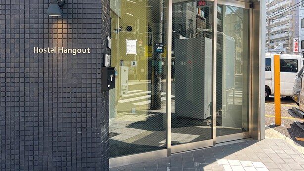 A hostel entrance with a glass door, a tiled wall, and a sign indicating 'Hostel Hangout'. - Hotel Hangout - Hostel (Osaka)
