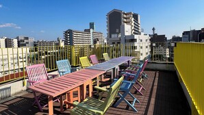 A rooftop terrace with a long table and colorful chairs, overlooking a cityscape with modern buildings. - Hotel Hangout - Hostel (Osaka)