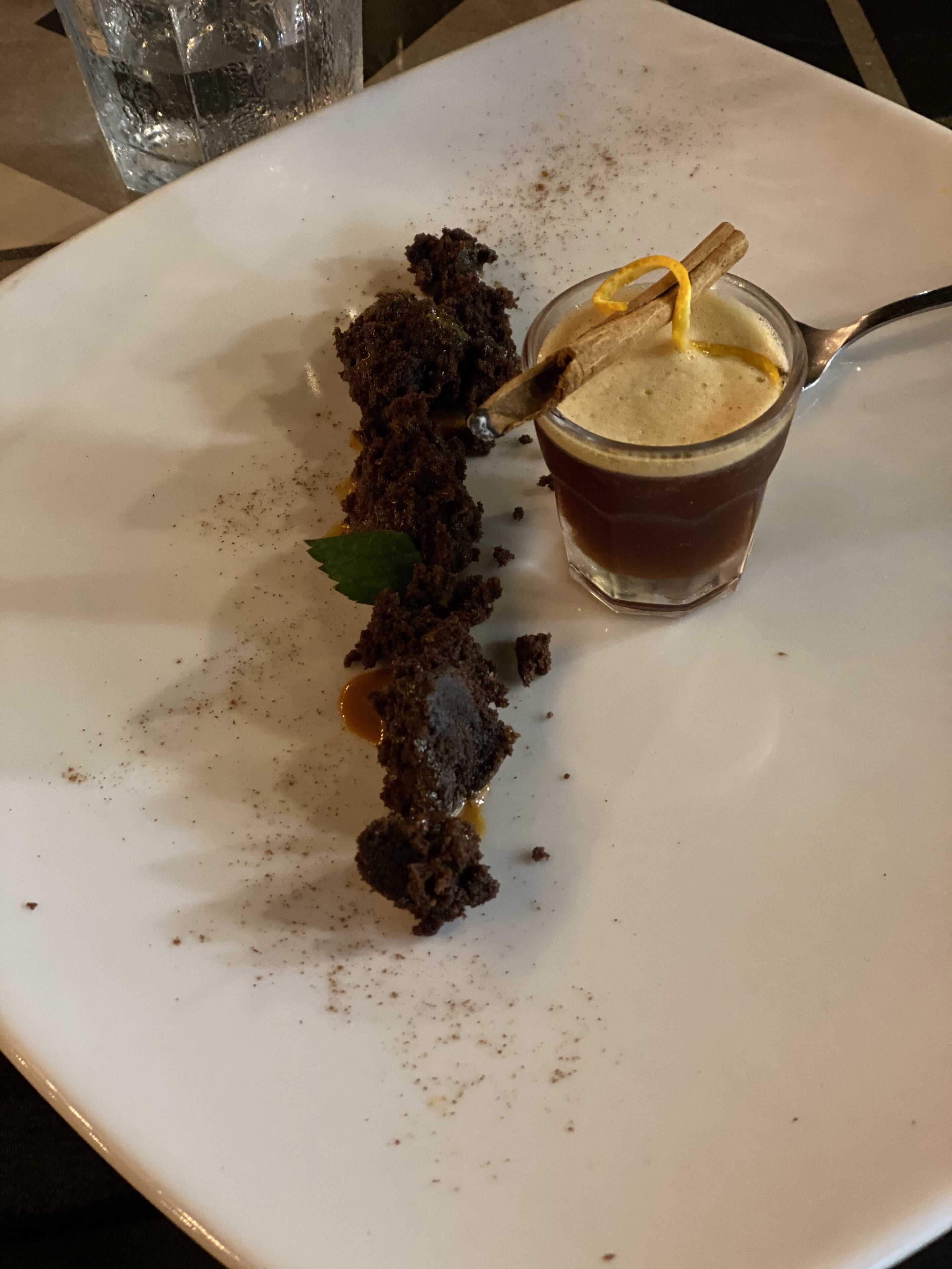 Een dessert op een wit bord, met een glas bruine vloeistof en een kaneelstokje.
