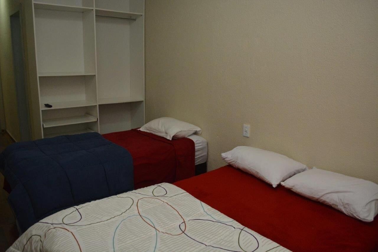 A room with two beds, one with a red bedspread and the other with a blue bedspread. There is a white wardrobe with open shelves to the left.