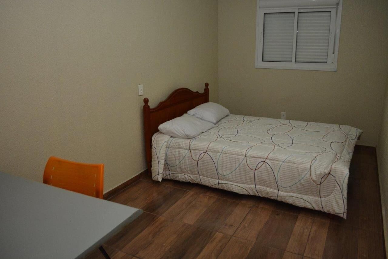 A small, neatly arranged bedroom with a single bed, a window with blinds, a wooden headboard, and a table with an orange chair.