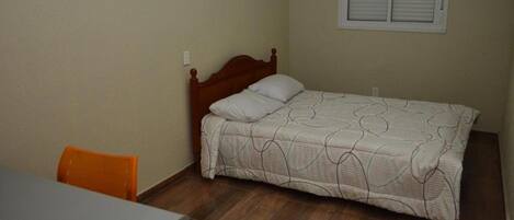 Un dormitorio pequeño y ordenado con una cama individual, una ventana con persianas, un cabecero de madera y una mesa con una silla naranja.