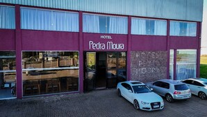 A hotel named 'Hotel Pedra Moura' with a purple facade, large windows, and a parking area with several cars.