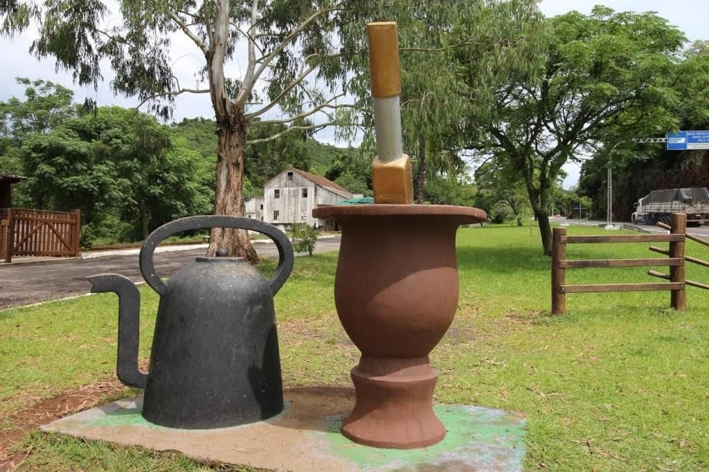 A black metal kettle and a terracotta pot with a handle, placed on a grassy area with a wooden fence and trees in the background.