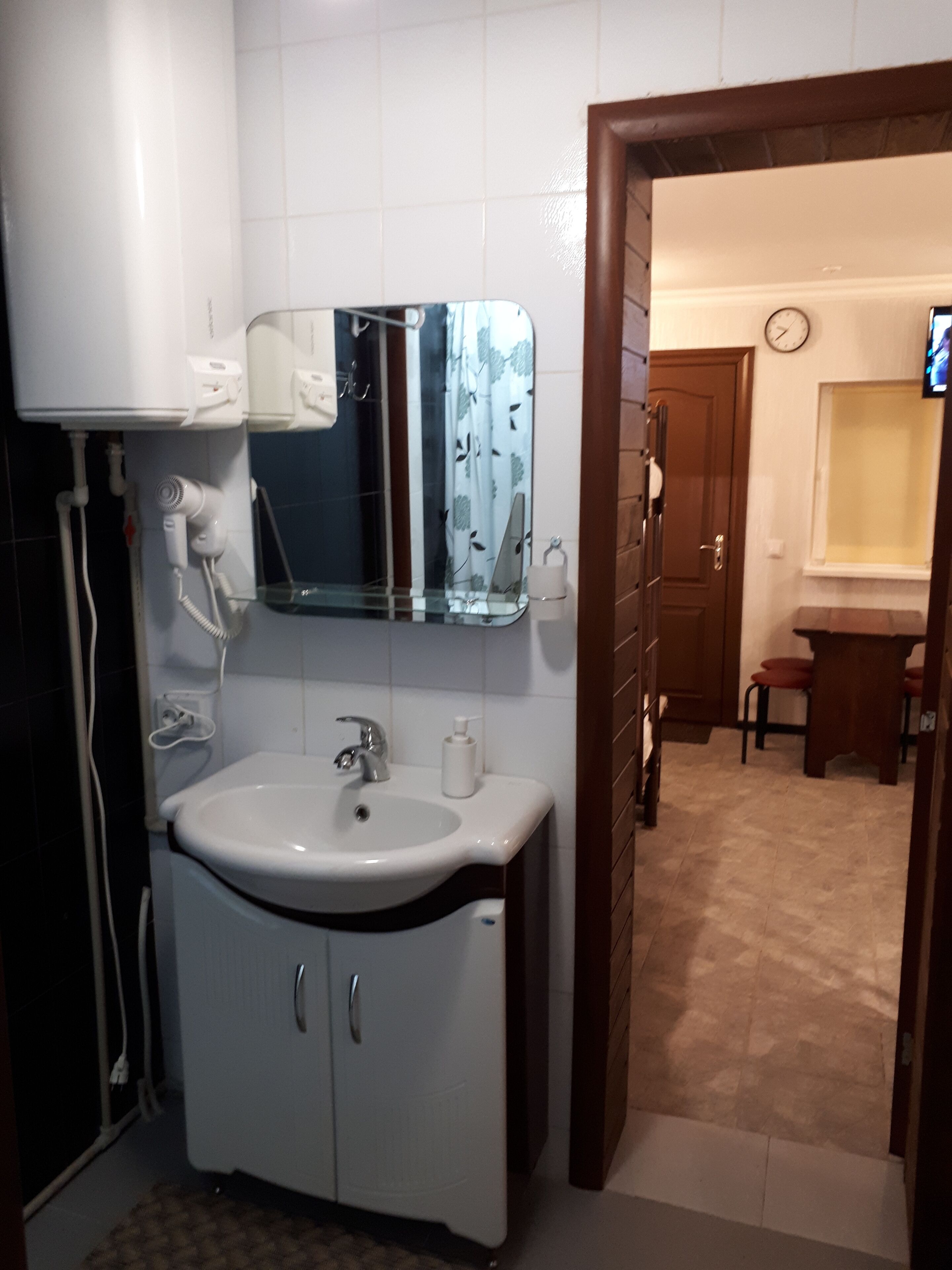 A bathroom with a white sink, a mirror, and a toilet. There is a clock on the wall and a TV in the corner.