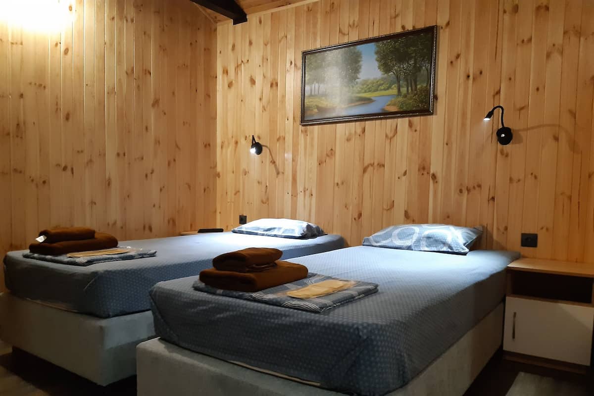 Ein Zimmer mit zwei Betten, beide mit blauer Bettdecke und braunem Handtuch. Darüber hängt ein Bild an der Wand, und rechts steht ein kleiner Tisch mit einer Lampe.