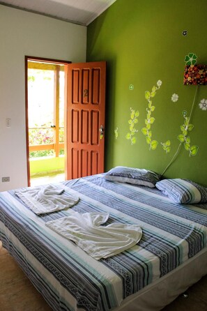 A bedroom with a bed, a green wall with floral decals, a wooden door, and a view of a garden through an open door.