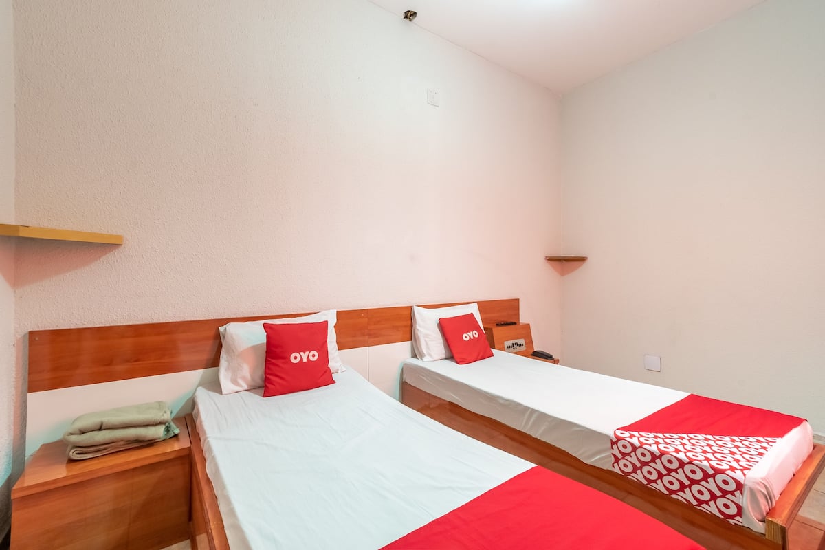 Een hotelkamer met twee enkele bedden, elk voorzien van een houten hoofdeinde en een rood-witte sprei. Er is een houten nachtkastje met een opgevouwen handdoek erop, en een klein plankje boven de bedden.