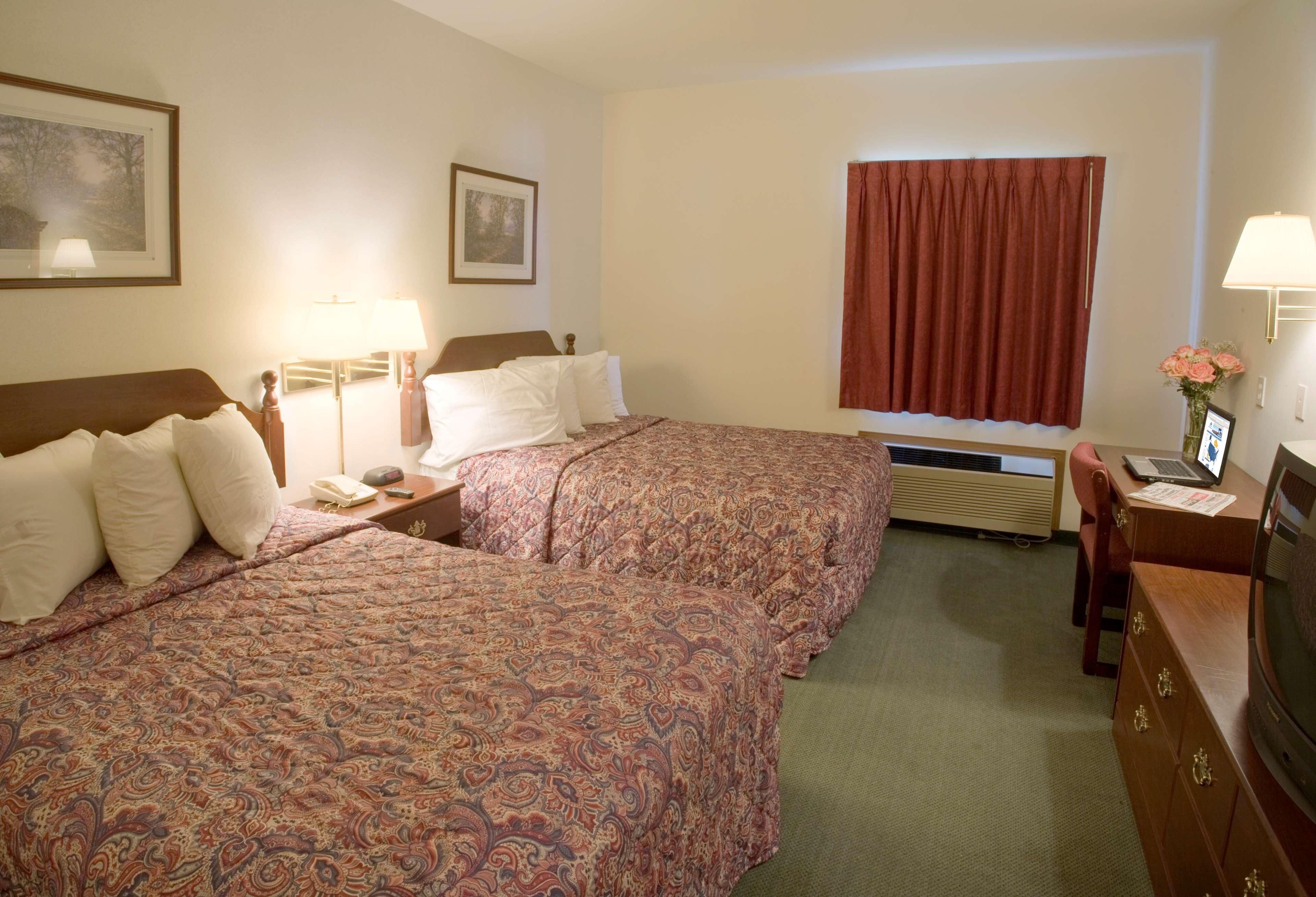 A hotel room with two beds, a desk with a laptop, a TV, a window with curtains, and a framed picture on the wall.