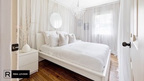 Une chambre à coucher avec un lit blanc, une table de chevet, un miroir et des rideaux en voile.