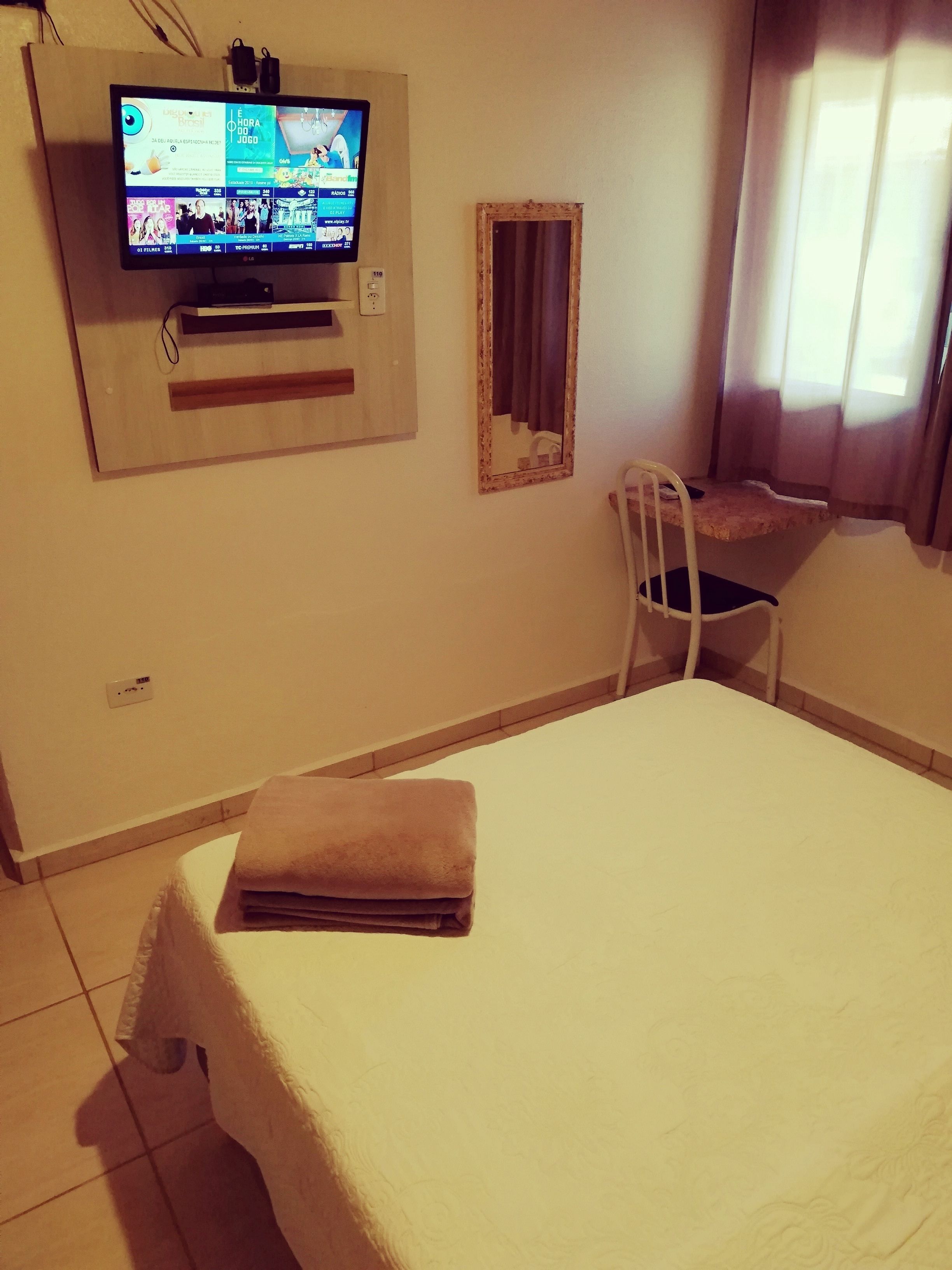 Habitación de hotel con una cama, una silla, un televisor fijado a la pared y una ventana con cortinas.