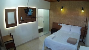 Habitación de hotel con cama, escritorio, silla, televisor montado en la pared y baño visible a través de una puerta de vidrio.