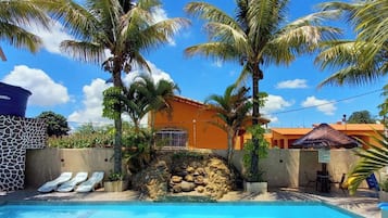 Un área de piscina con sillones de descanso, palmeras y un edificio con pared de piedra.