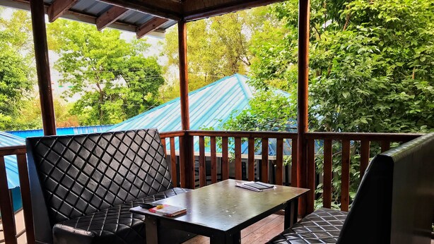 A covered outdoor seating area with two cushioned chairs and a table, surrounded by wooden railings and greenery. - Park Hostel Osh (Osh)