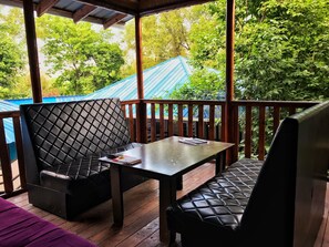 A covered outdoor seating area with two cushioned chairs and a table, surrounded by wooden railings and greenery. - Park Hostel Osh (Osh)