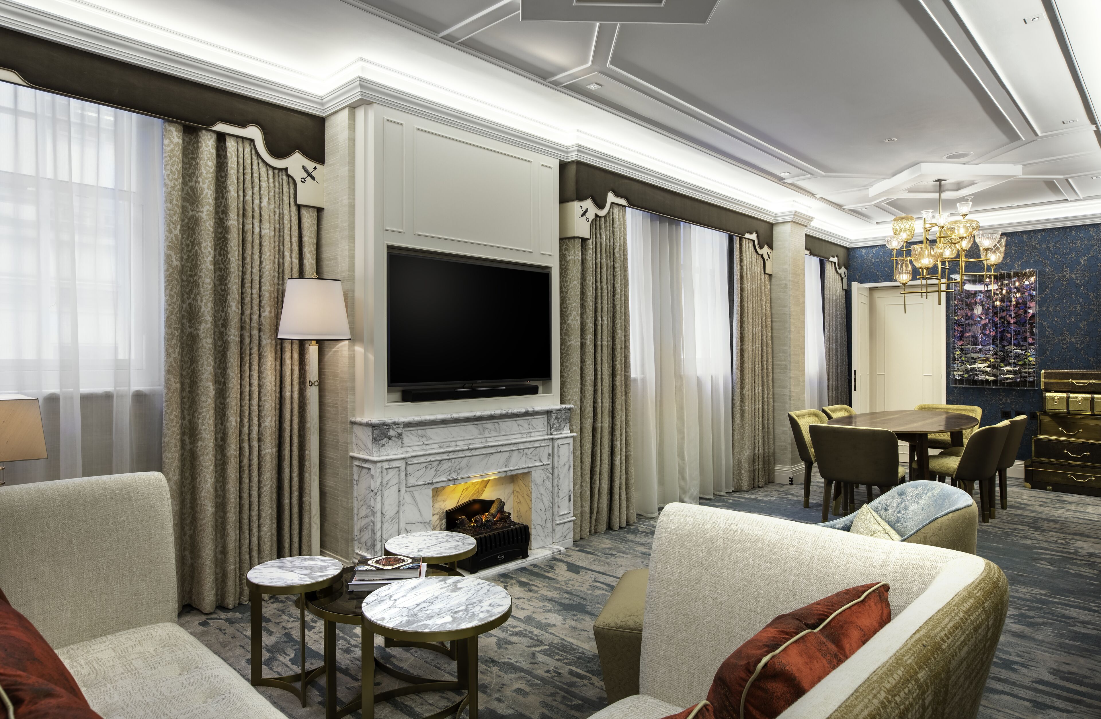 A luxurious hotel room with a fireplace, a flat-screen TV, a dining area, and a chandelier.