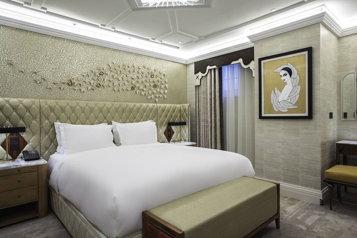 Une chambre spacieuse avec un grand lit, une tête de lit capitonnée, une table de chevet avec une lampe et une œuvre d’art encadrée au mur.