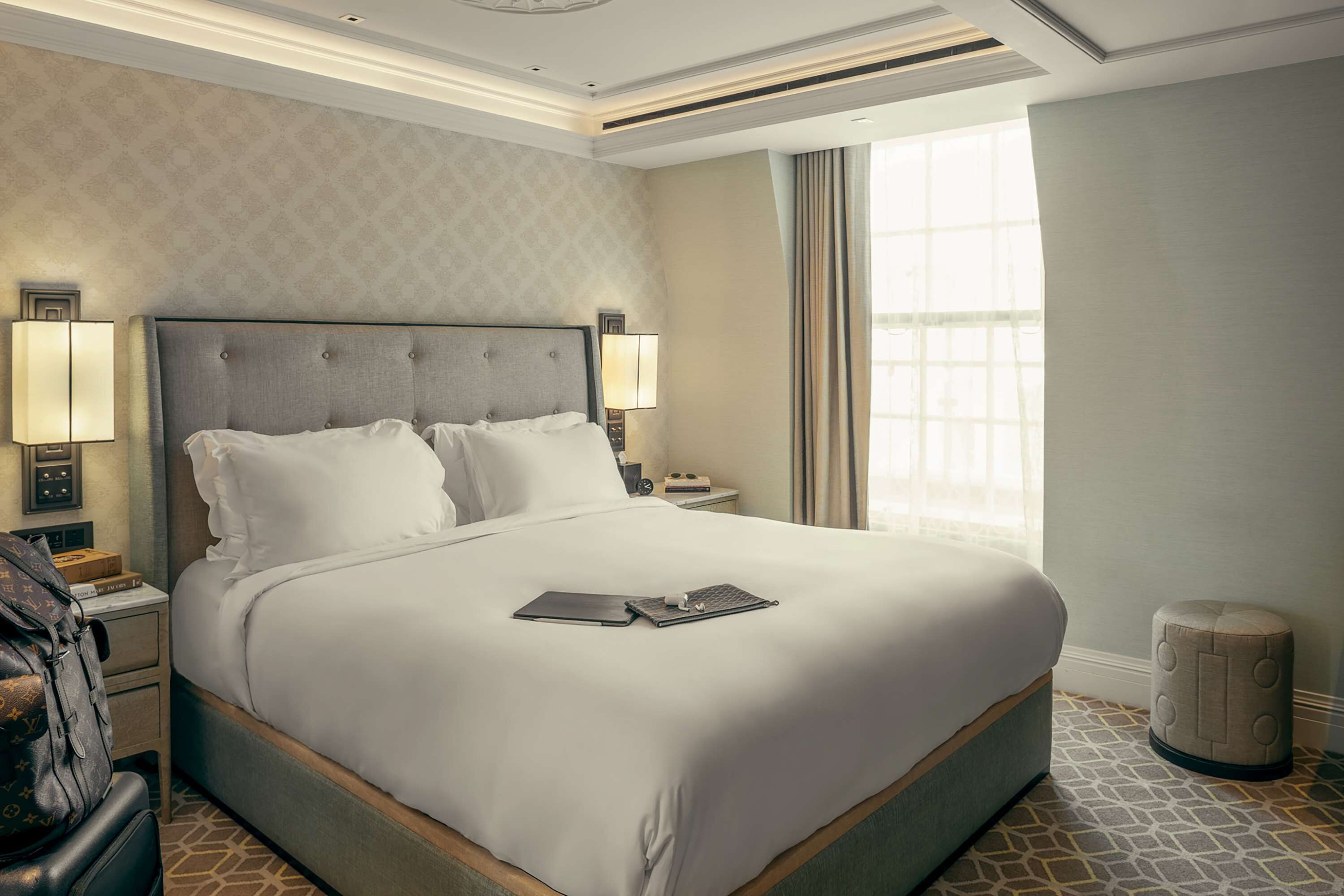 A hotel room with a large bed, two bedside lamps, a nightstand, a suitcase, and a window with curtains.