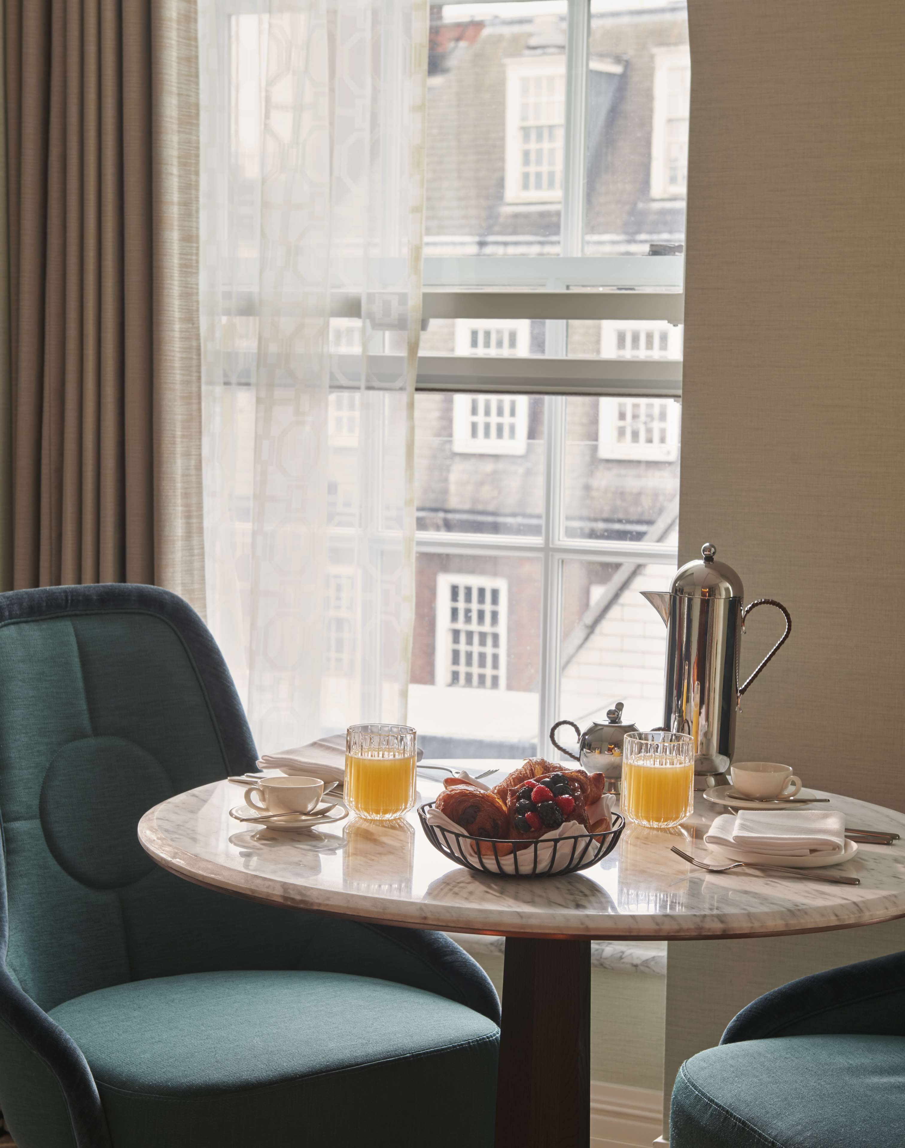 A breakfast table with croissants, a fruit bowl, a coffee pot, and a glass of orange juice.