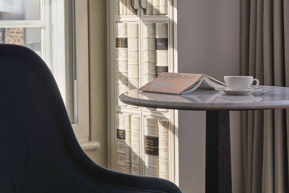 Une pièce avec une fenêtre, une chaise, une table sur laquelle se trouvent un livre et une tasse, et une bibliothèque.