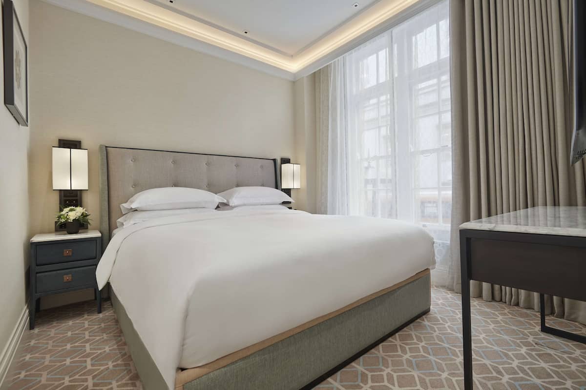 Une chambre d’hôtel avec un grand lit, des tables de chevet avec des lampes, une petite table et une vue sur l’extérieur à travers une fenêtre avec des rideaux.
